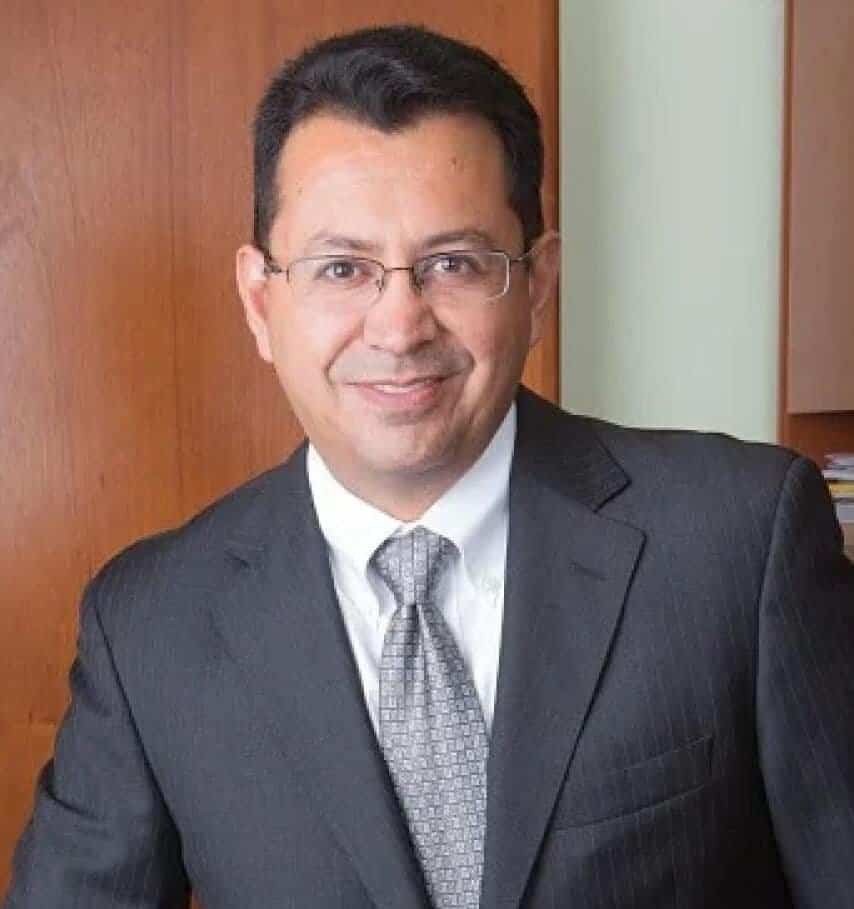 Man in a dark suit and patterned tie stands in an office setting with a wooden background. He has short black hair and is wearing glasses, smiling at the camera.